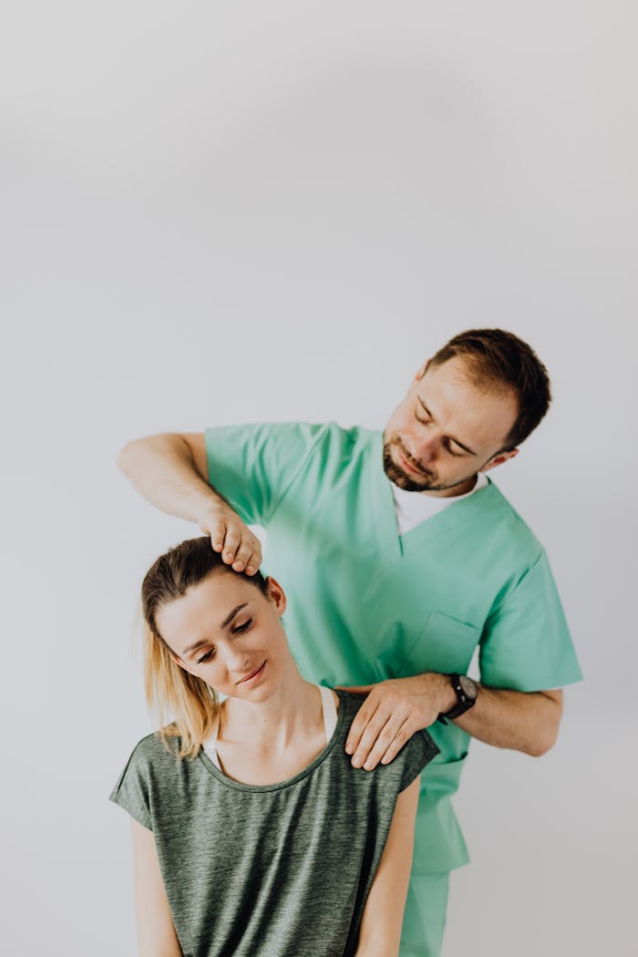 Positive professional male massage therapist wearing green medical uniform while standing behind positive woman and doing rehabilitation massage on neck muscles against gray background